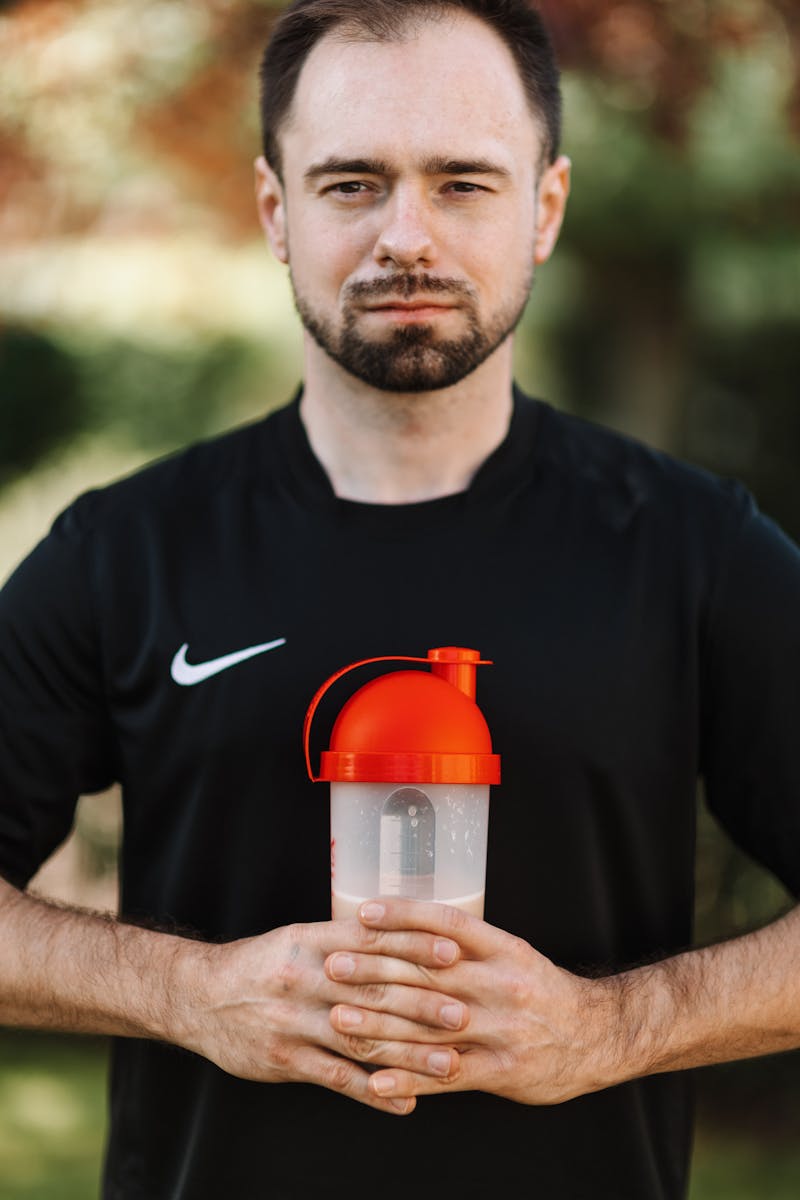 Bearded man in black shirt holding a protein shaker, standing outdoors. Fitness and health concept.